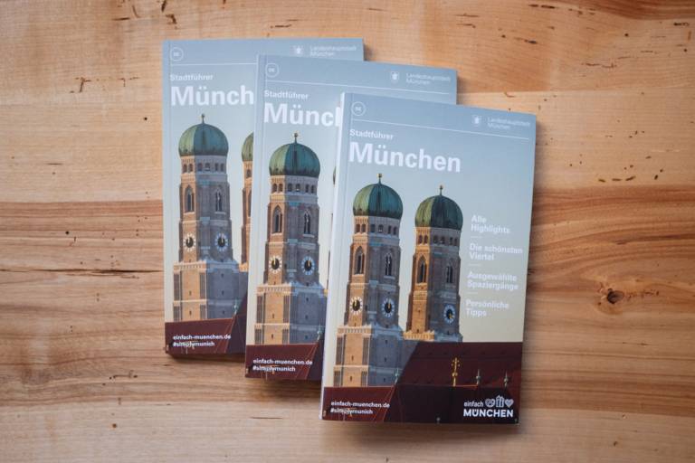 Three copies of the official Munich city guide are slightly stacked on top of each other so that the image of the Frauenkirche remains visible on each one.